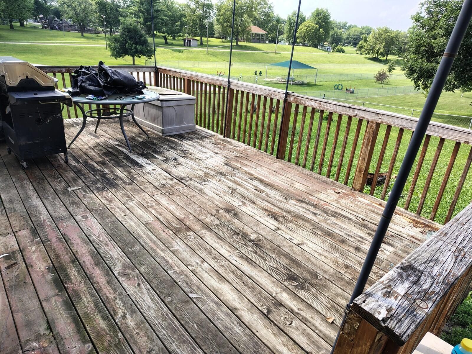Grungy wooden deck with grill, table, and railing overlooking a grassy park on a cloudy day.