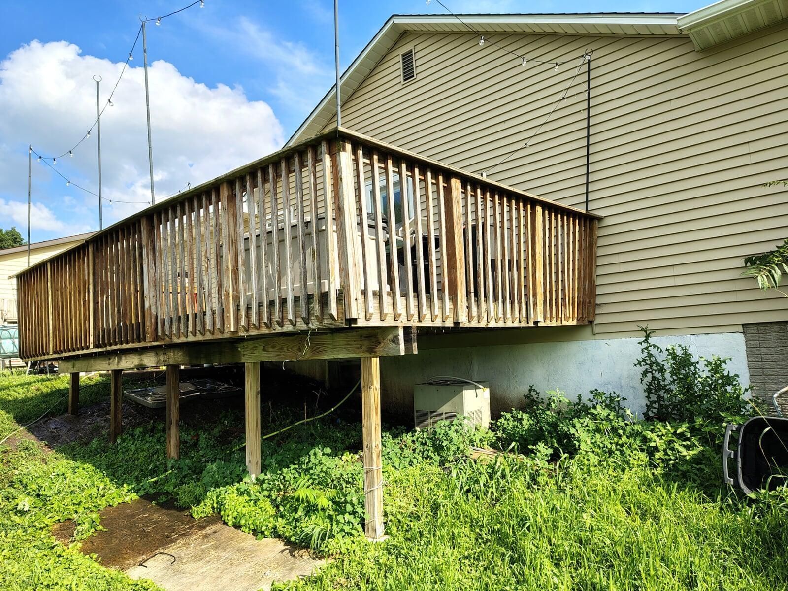 A wooden deck attached to a light beige building. The deck is elevated on wooden supports, surrounded by tall green grass.