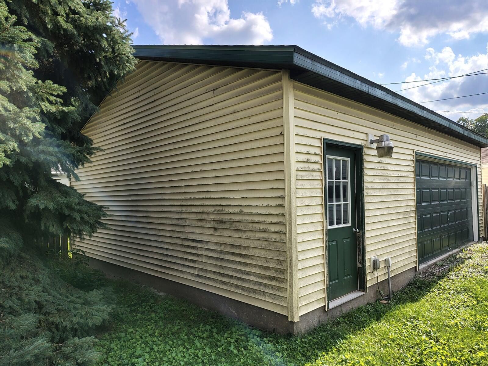 Garage with yellow siding, green door and garage door, under a cloudy sky. The structure is on a grassy area with a nearby tree.