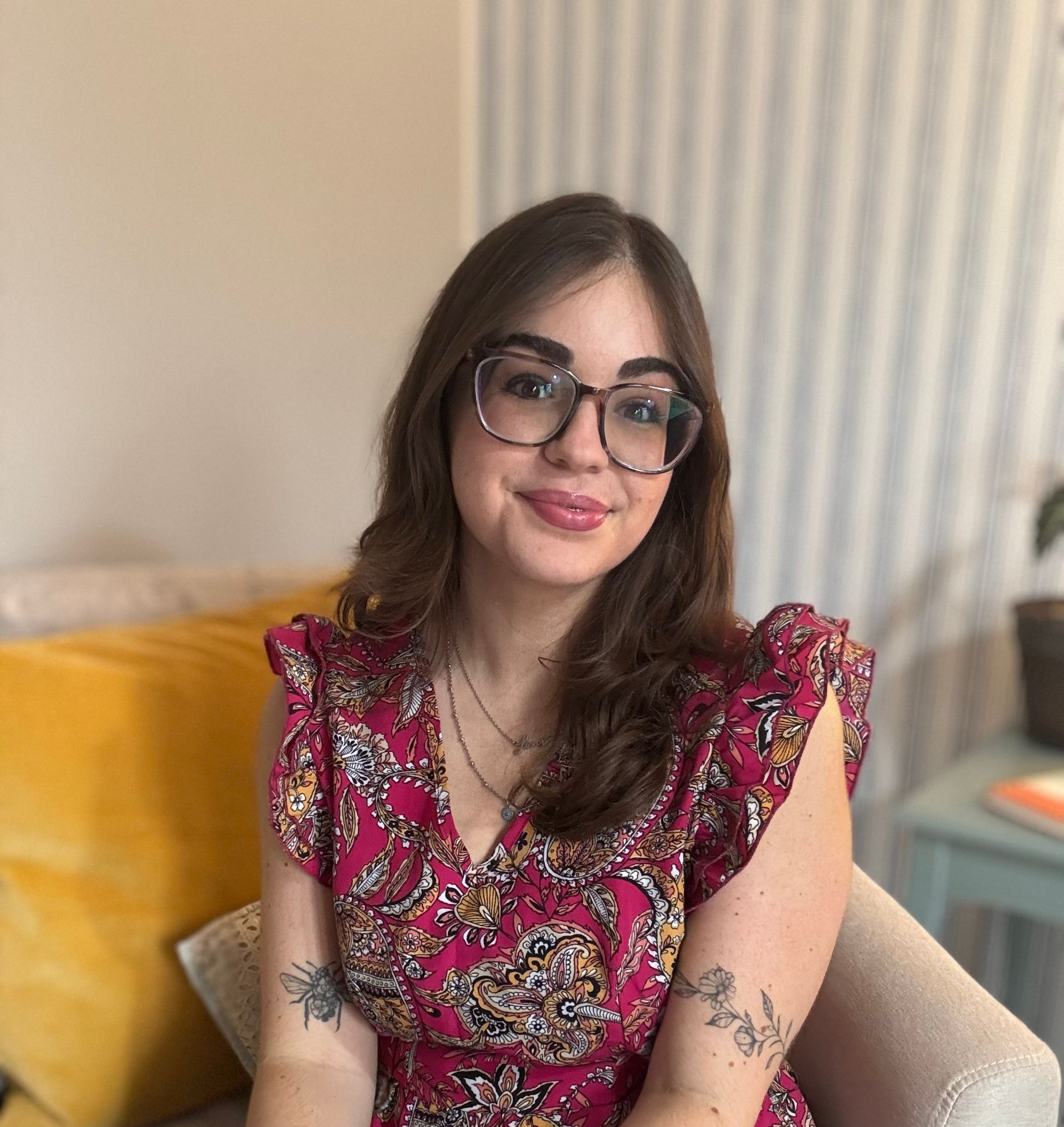 Woman with glasses, wearing a red patterned dress, seated indoors, smiling at the camera.