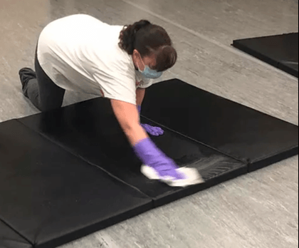 A woman wearing a mask and gloves is cleaning a black mat.