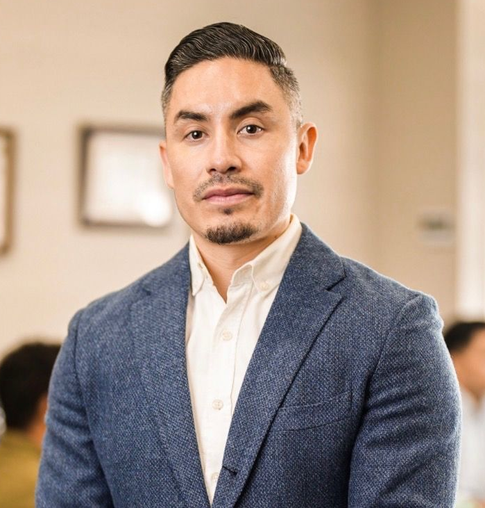 Man in blue blazer, hands in pockets, looking at camera in office setting.