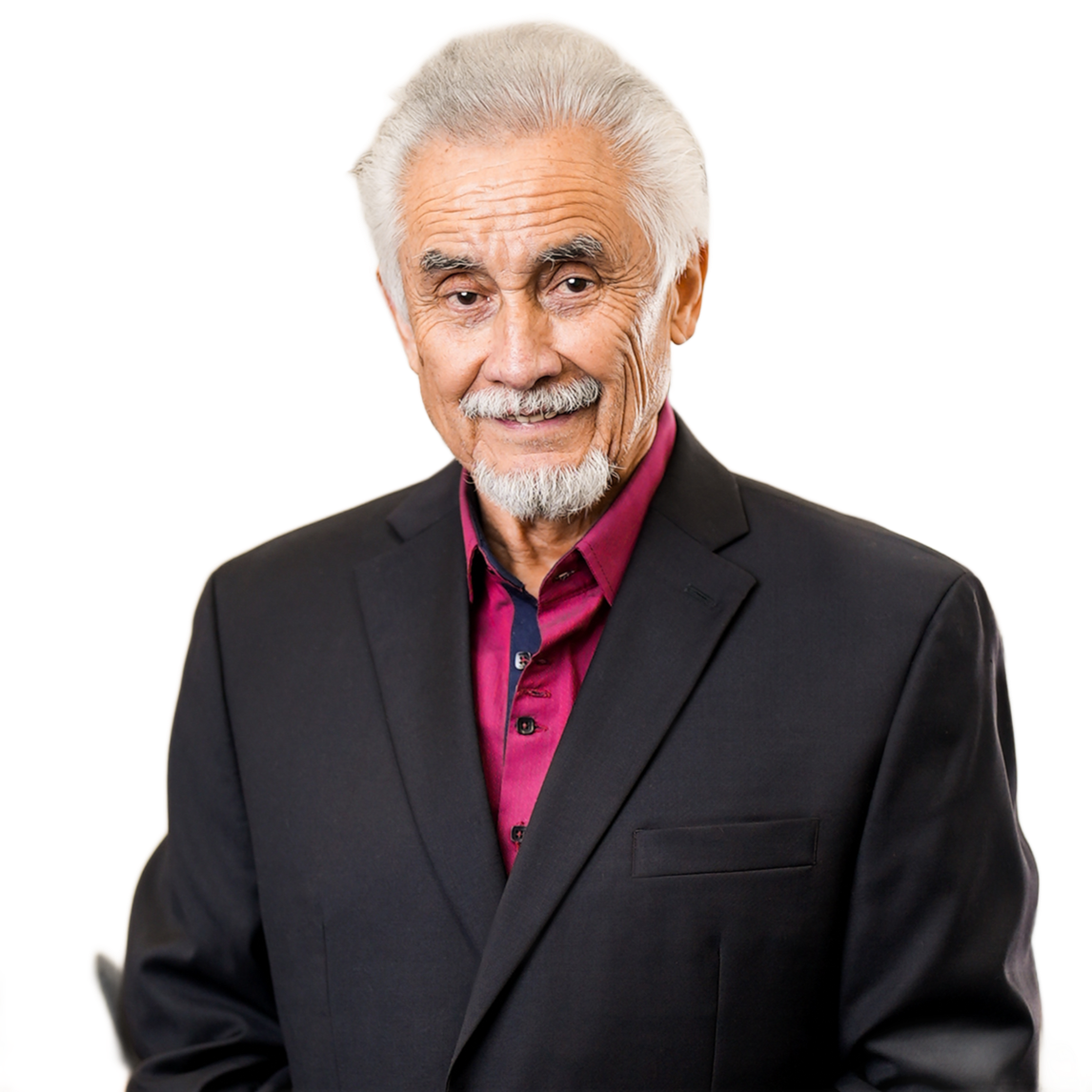 Man in a suit smiles, standing indoors; background blurred with frames and office equipment.