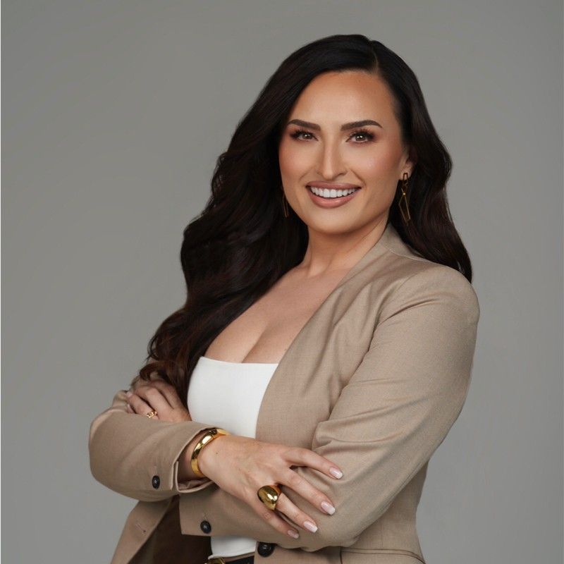 Woman in beige blazer, arms crossed, smiling, with gold jewelry, against a gray backdrop.
