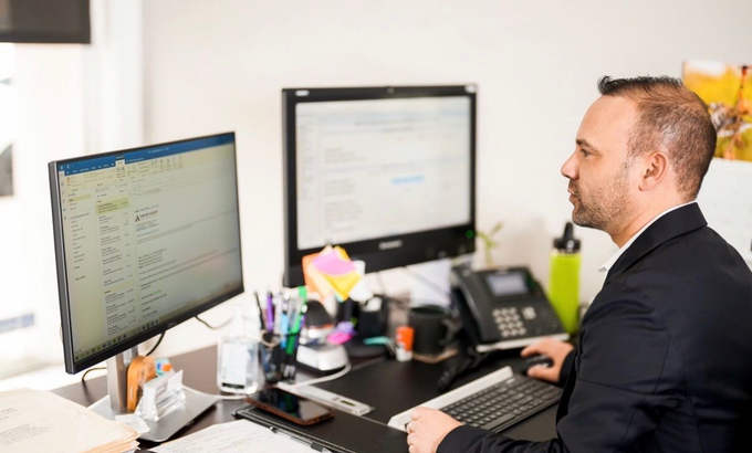 Man in suit working at a desk with two computer monitors, phone, and office supplies.