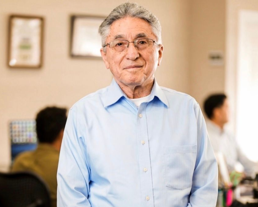 Man in a light blue shirt and glasses, smiling in an office setting, framed pictures on wall, and employees in background.