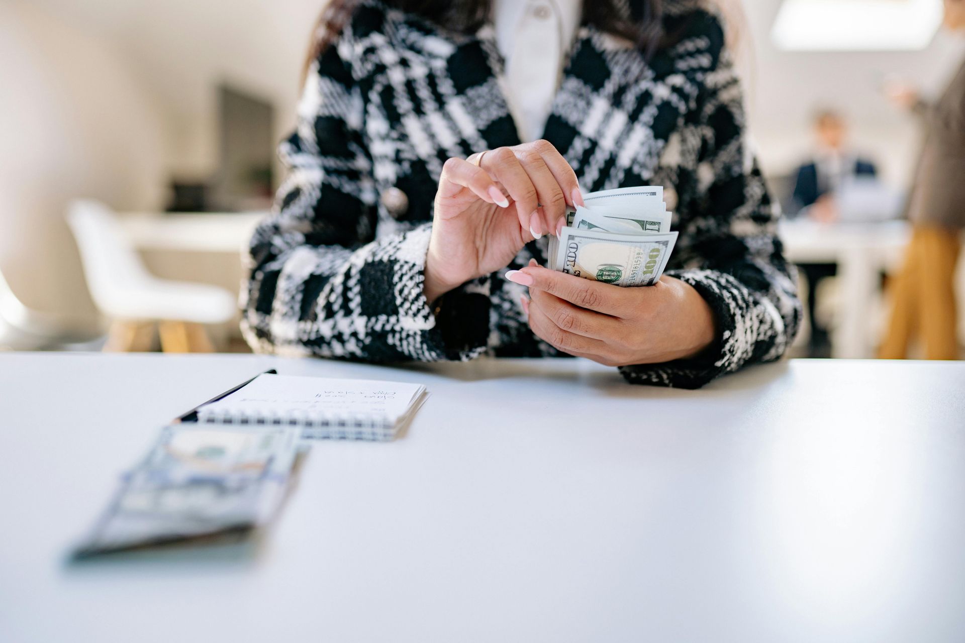 Person counting money at a white table, small notepad nearby, with a plaid jacket on.