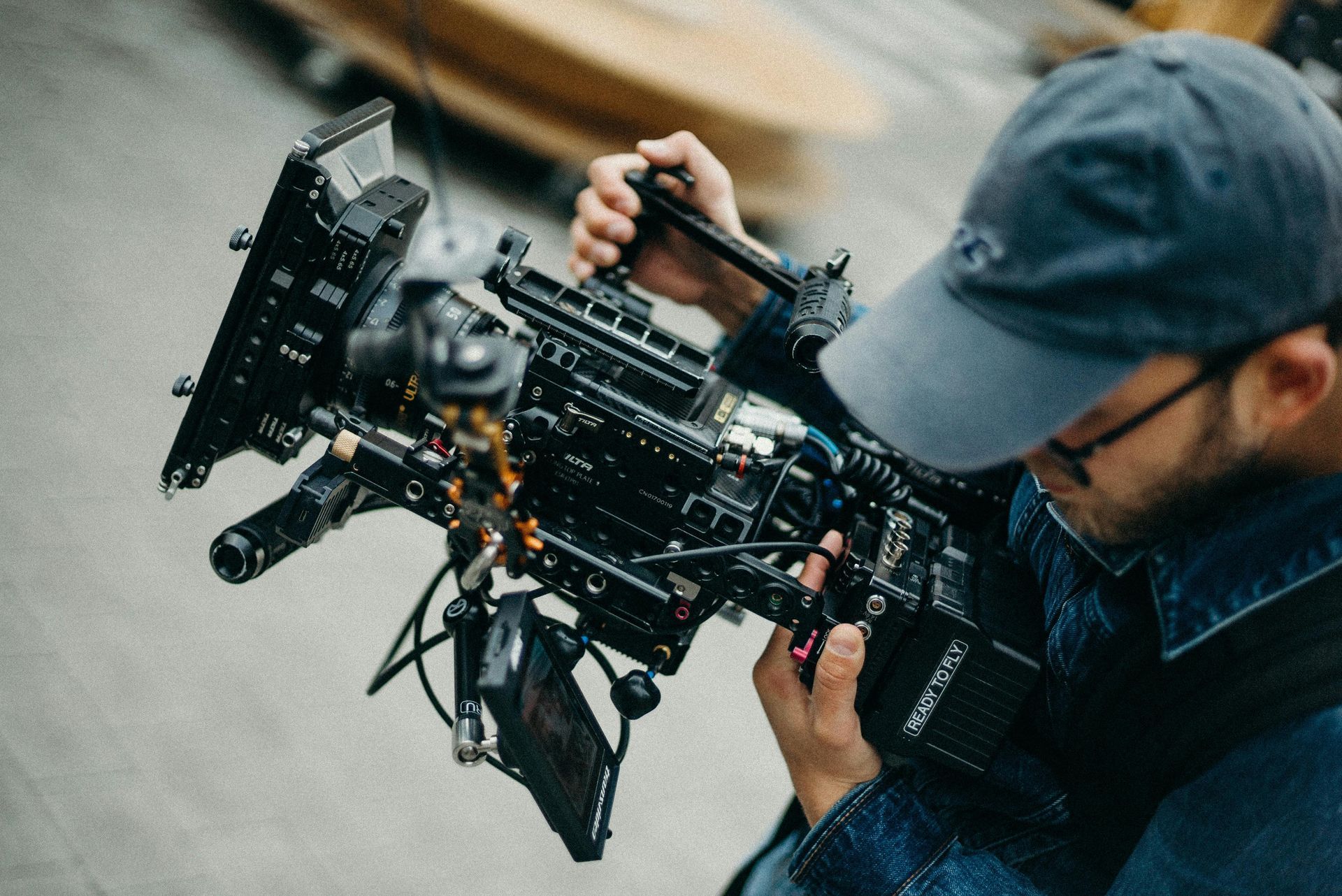 Person holding a large, black camera with accessories, wearing a blue cap and glasses.