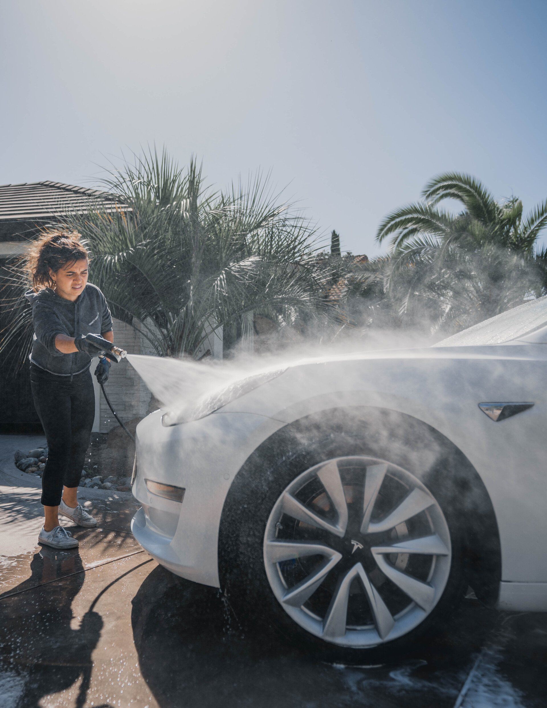Young person washing a white Tesla car with soapy water outdoors on a sunny day.