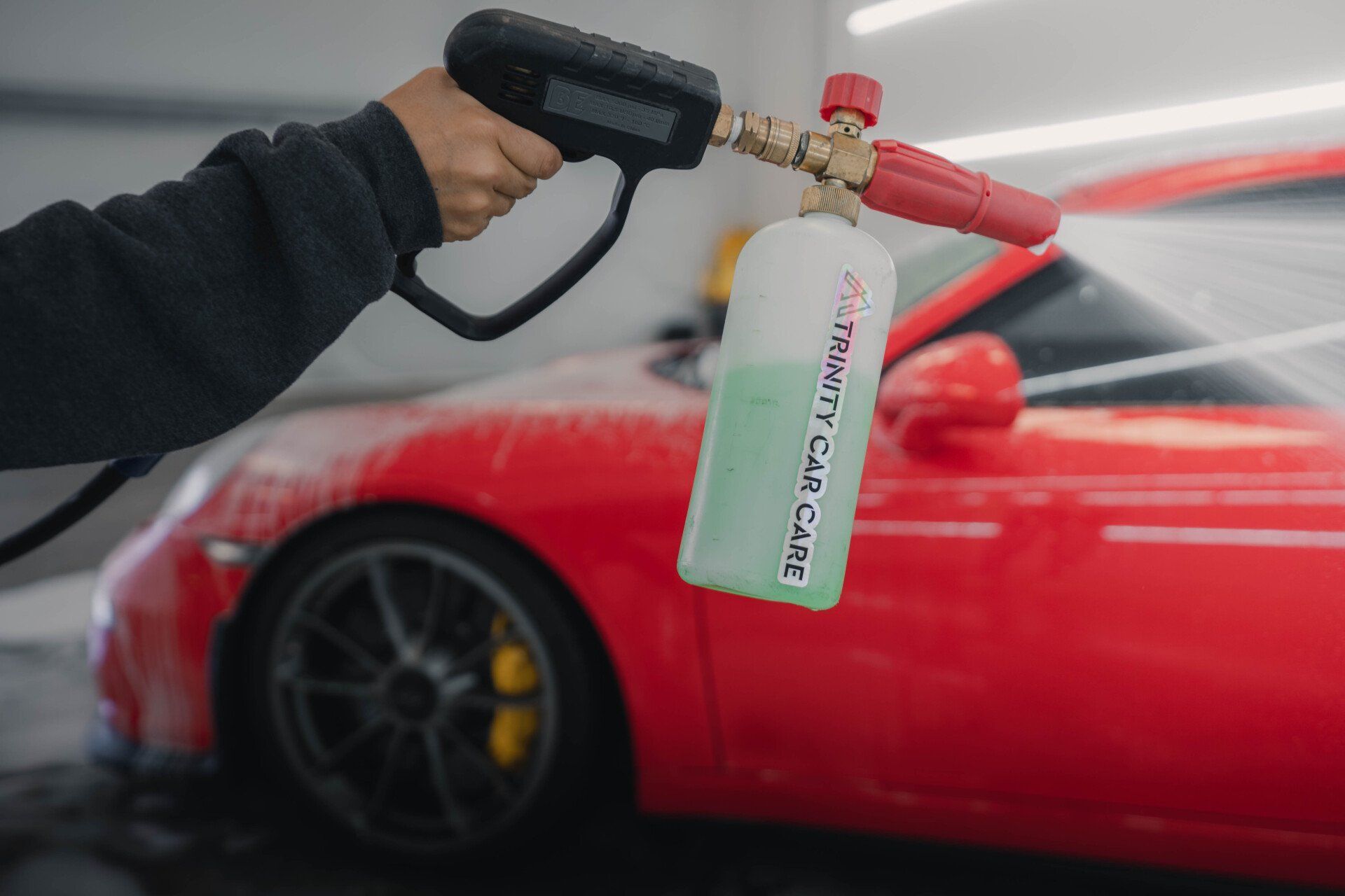 A person sprays foam onto a red car with a foam cannon in a car wash setting.