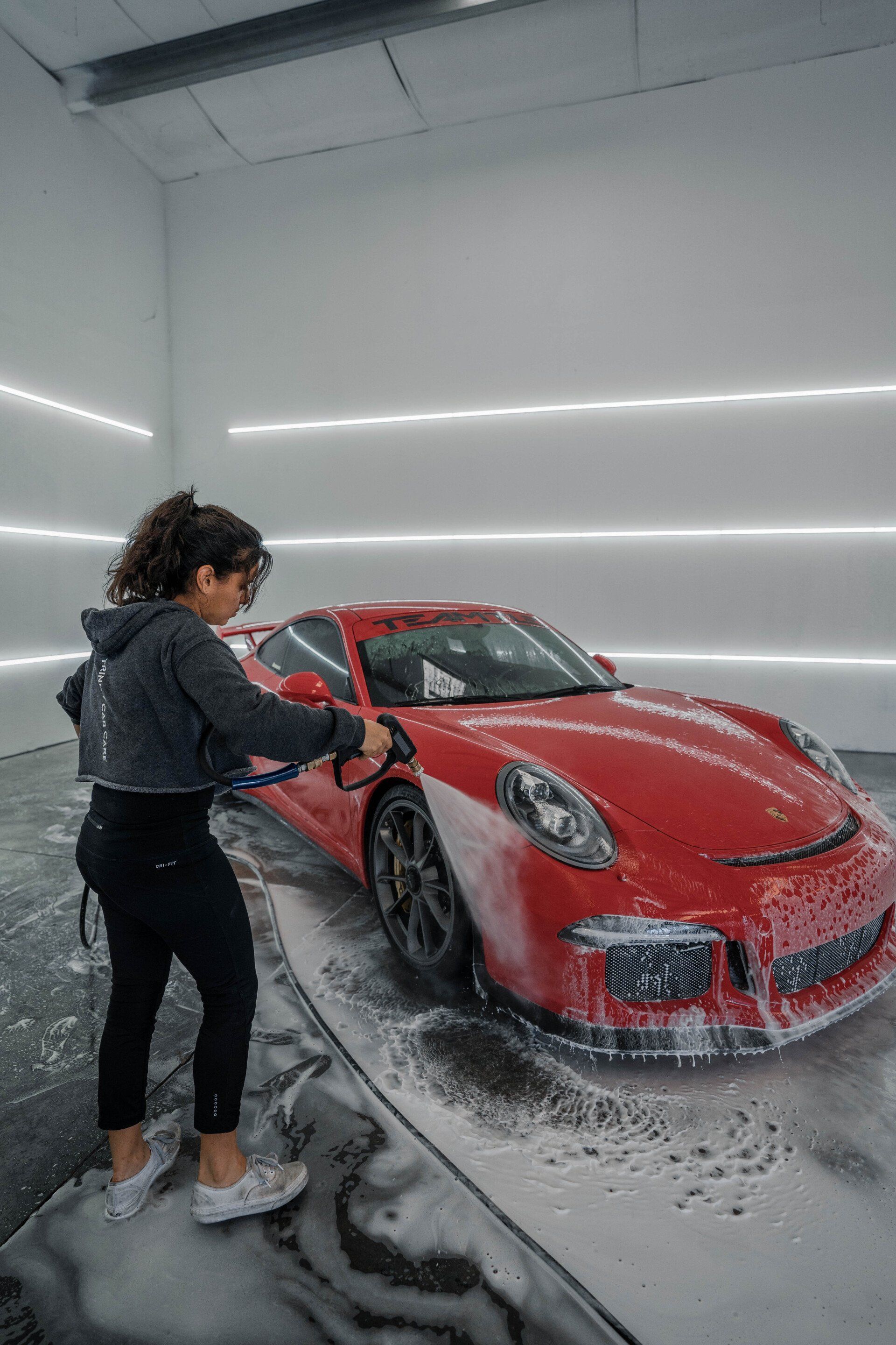 Person washing a red sports car with a foam spray in a brightly lit garage.