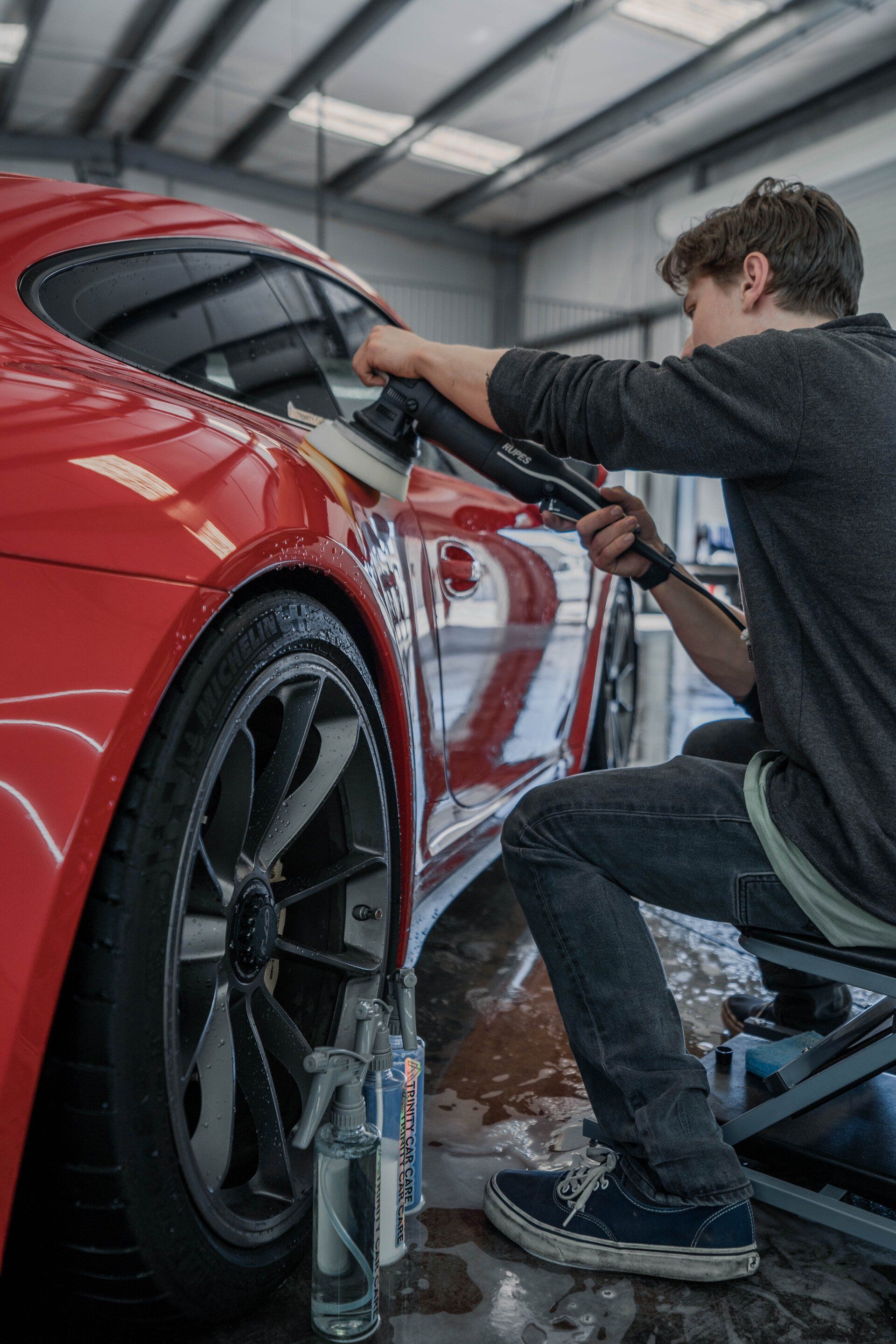 Person polishing a red car in a car wash. They are using a polisher and sitting on a stool.