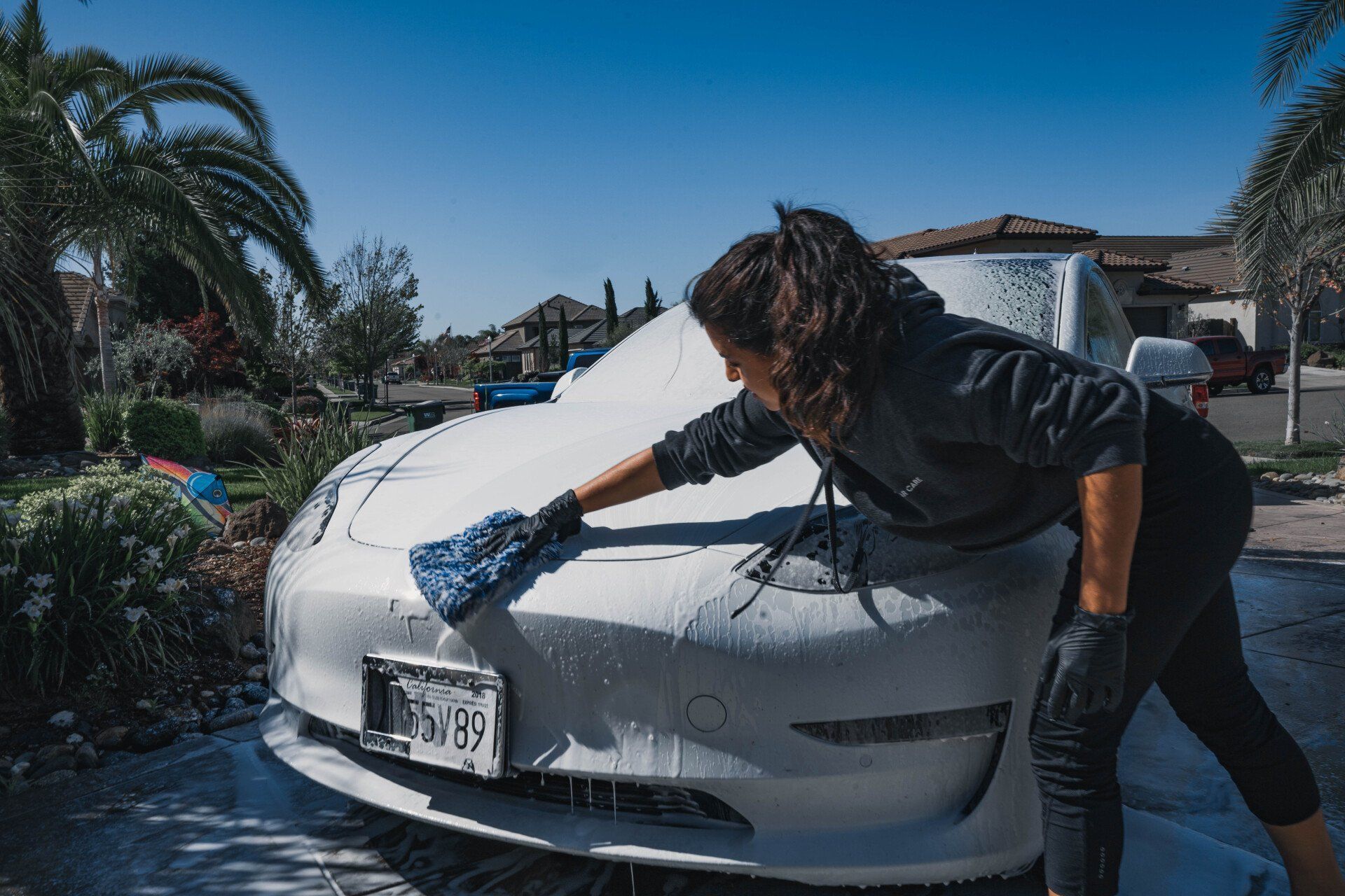 Woman washing a white car with foamy soap in a driveway on a sunny day.