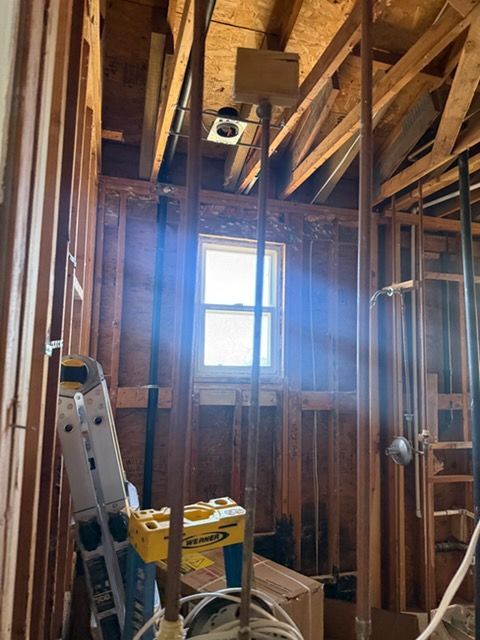 An unfinished room under construction with exposed wooden wall studs, ceiling joists, vertical pipes, and a window.