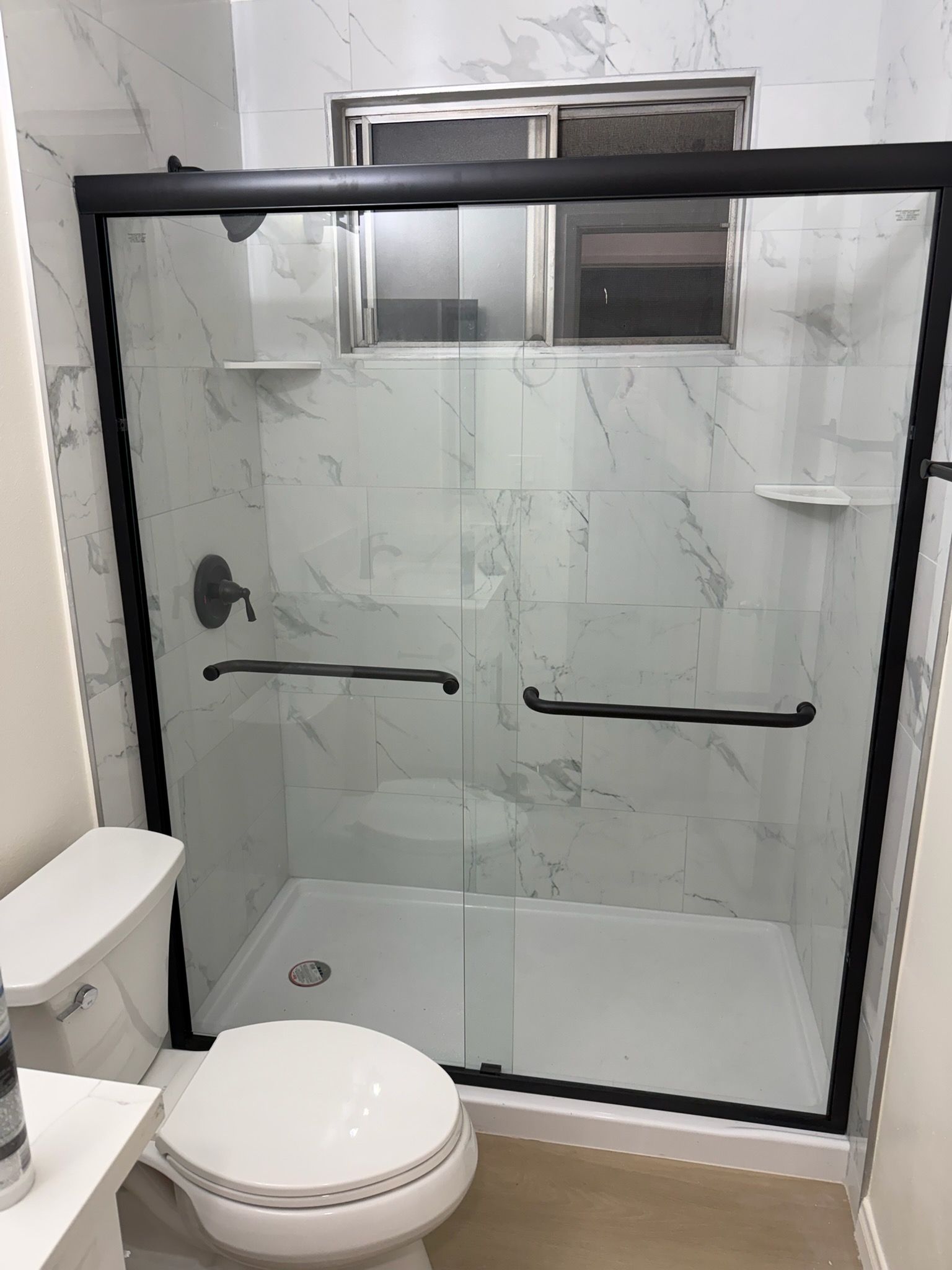 A bathroom shower with sliding glass doors, black frame, and white marble-patterned tile, positioned next to a toilet.
