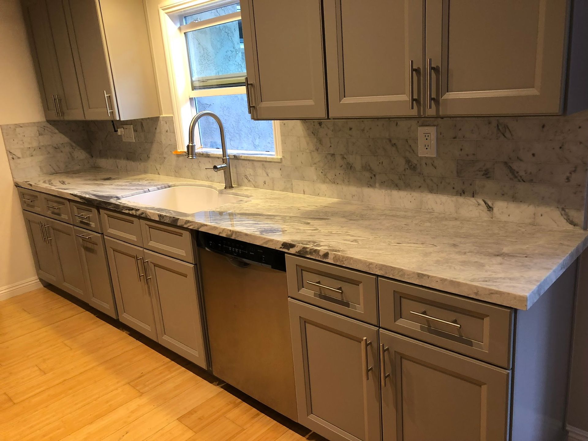 A kitchen with gray cabinets , marble counter tops , a sink and a dishwasher.