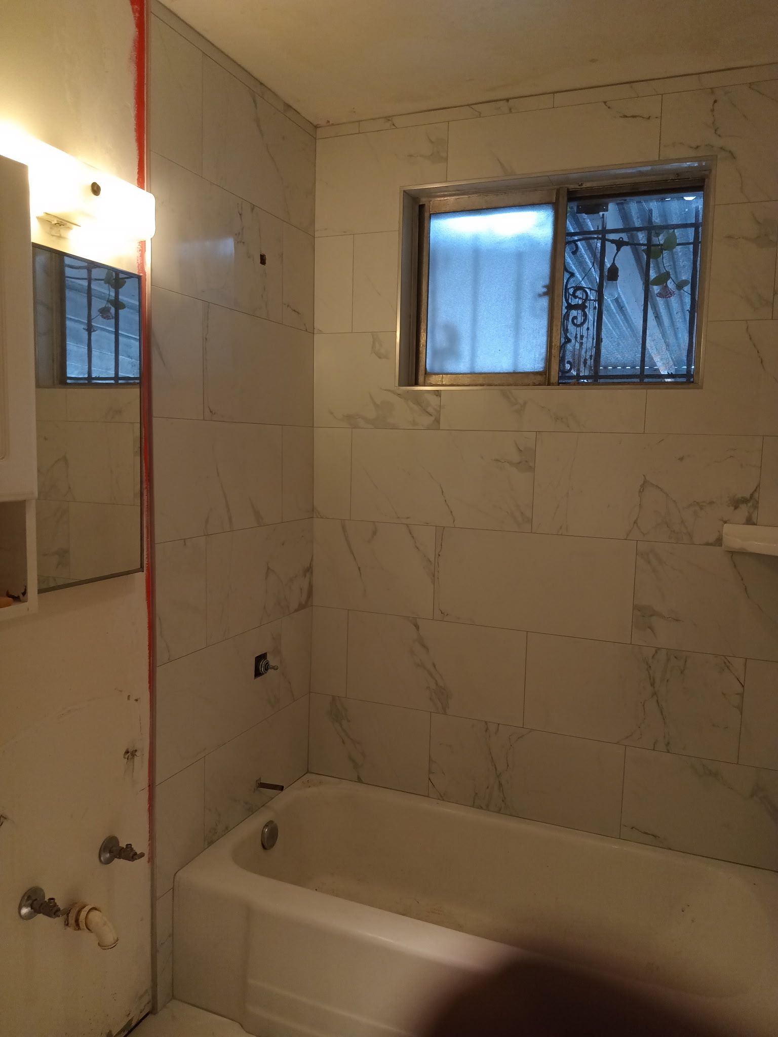 Bathroom with white marble-like tile, a bathtub, a window with security bars, and a partially visible mirror.
