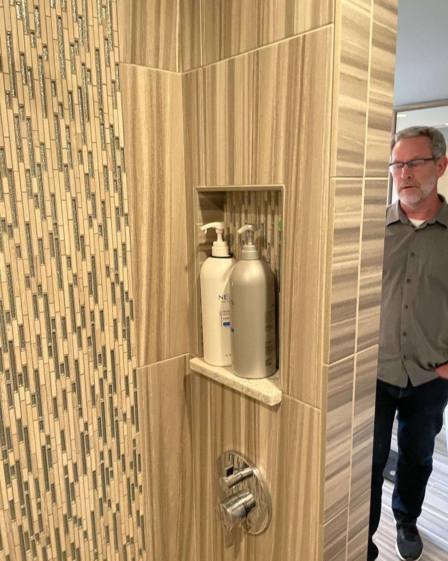 A man is standing in a bathroom next to a shower with two bottles of soap on a shelf.