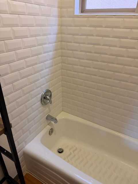 A white bathtub with subway-style wall tiles and chrome fixtures, positioned beneath a window in a bathroom.