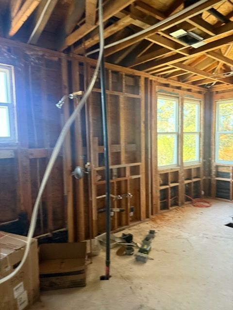 A room under construction with exposed wooden wall studs, ceiling joists, visible plumbing pipes, and two large windows.