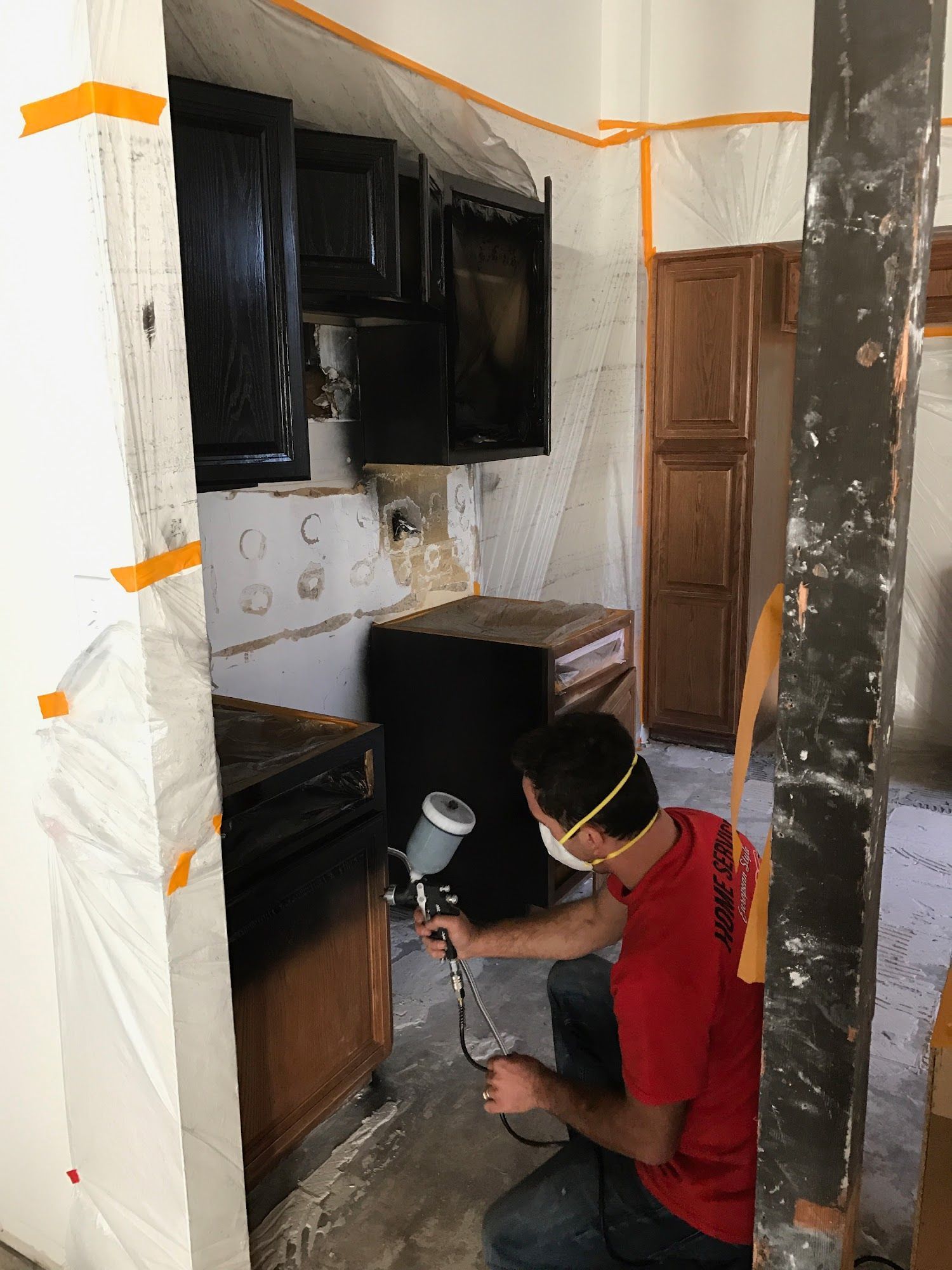 A man in a red shirt is spray painting a kitchen cabinet.