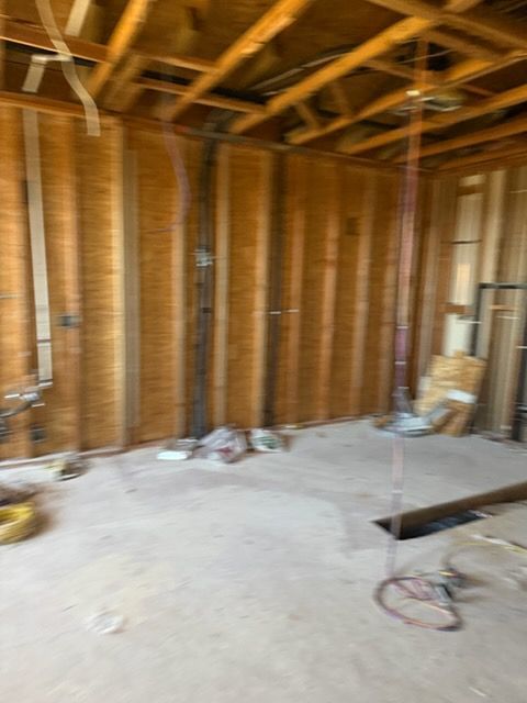 An interior view of a room under construction, featuring exposed wooden wall studs, ceiling joists, and a concrete floor.