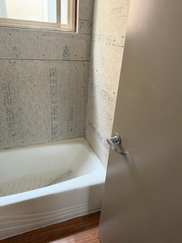A white bathtub in a bathroom with unfinished gray cement board walls and a light brown door in the foreground.