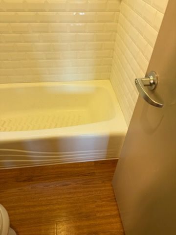A view of a white bathtub next to a closed door with a metal lever handle, set against a tiled wall and wood-look floor.
