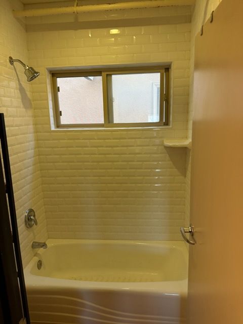 A white bathtub with a showerhead and subway tile surround beneath a sliding horizontal window.