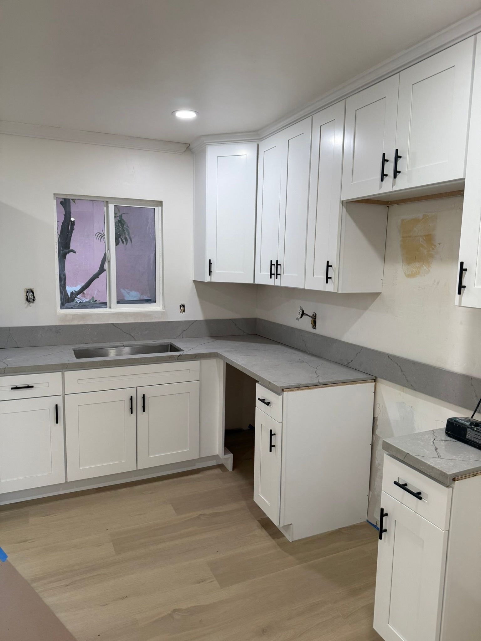 A kitchen with white cabinets, dark hardware, grey countertops, and light wood flooring under a single recessed light.