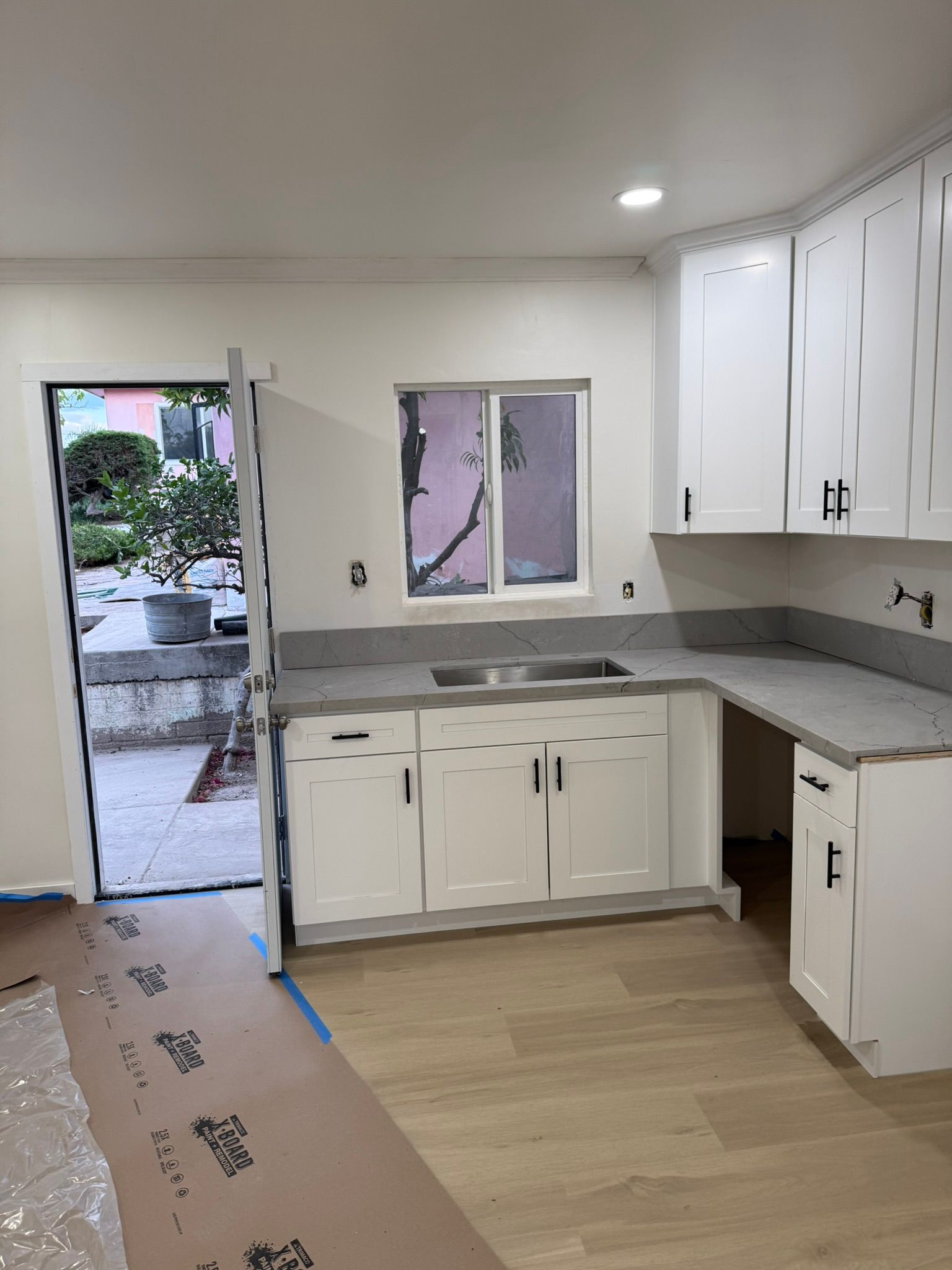 A kitchen under construction with white cabinets, gray countertops, and a doorway leading outside.