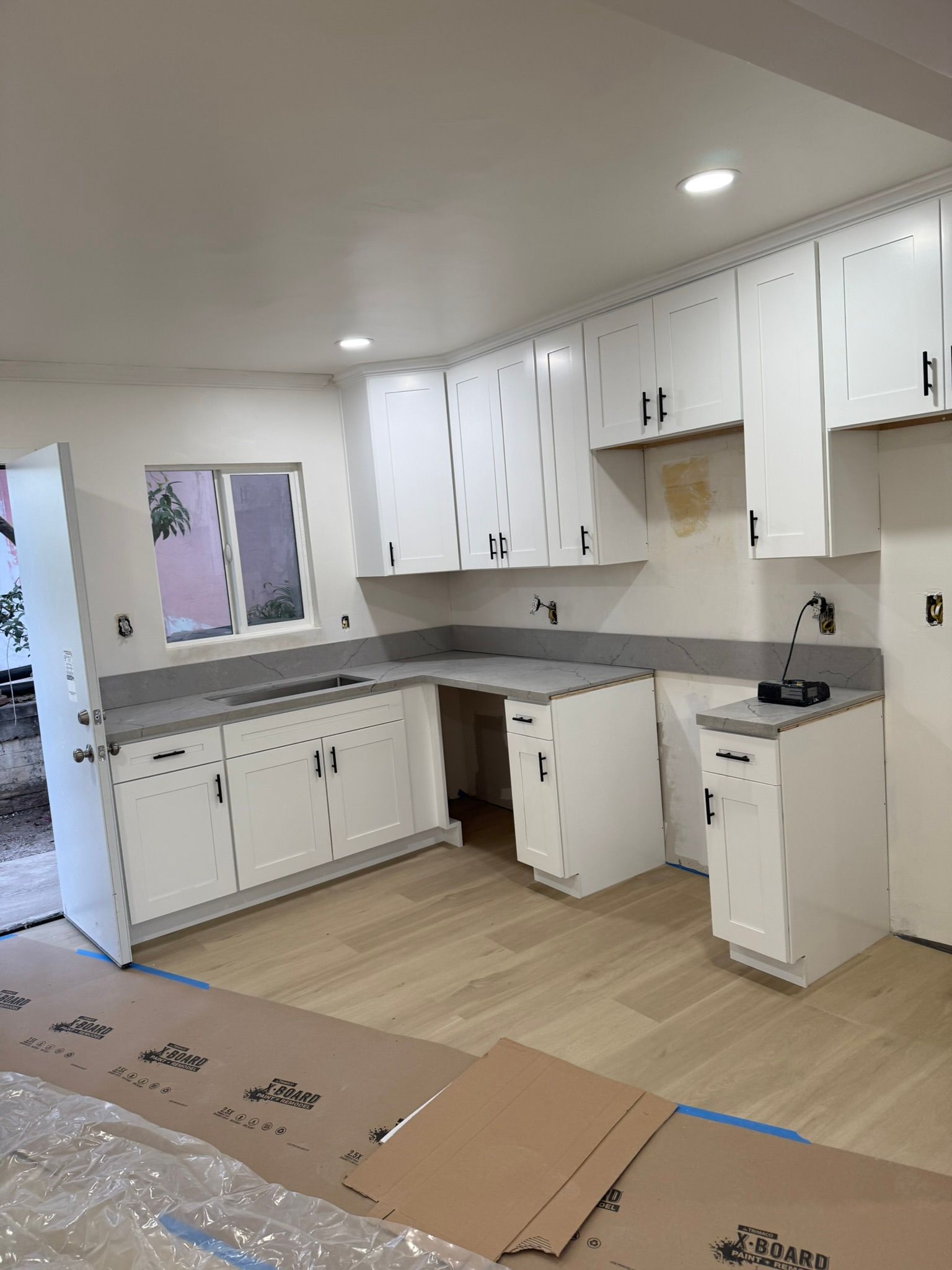 A white kitchen under renovation with new cabinets, light wood flooring, gray countertops, and protective floor paper.