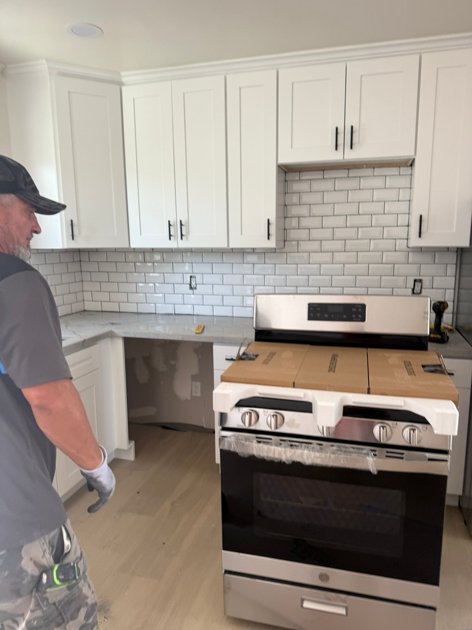 A worker in a gray shirt stands in a kitchen with new white cabinets, a subway tile backsplash, and a boxed-in stove.