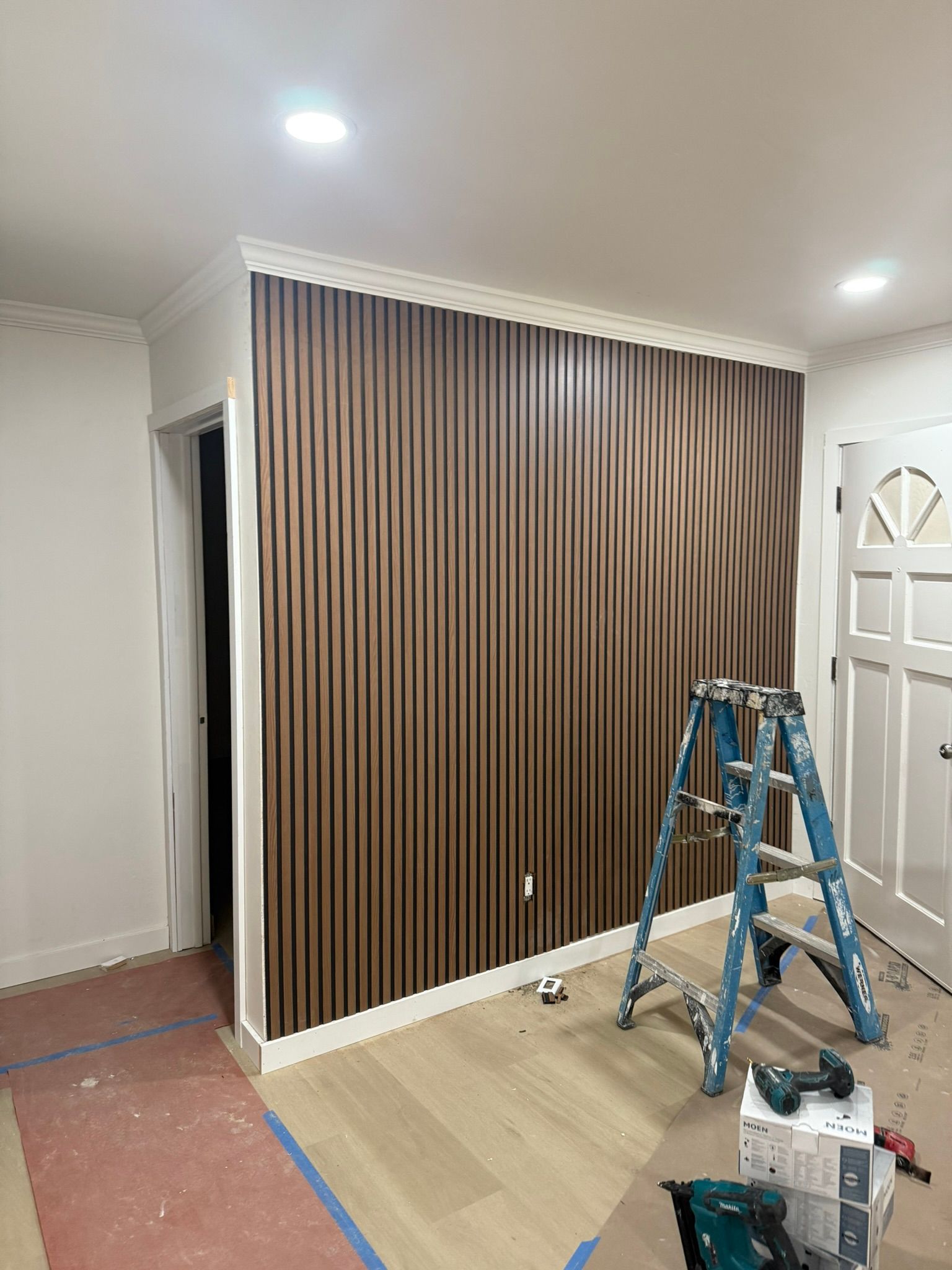 A wall featuring vertical brown wood slats, with a blue stepladder and tools on the floor in a room under construction.