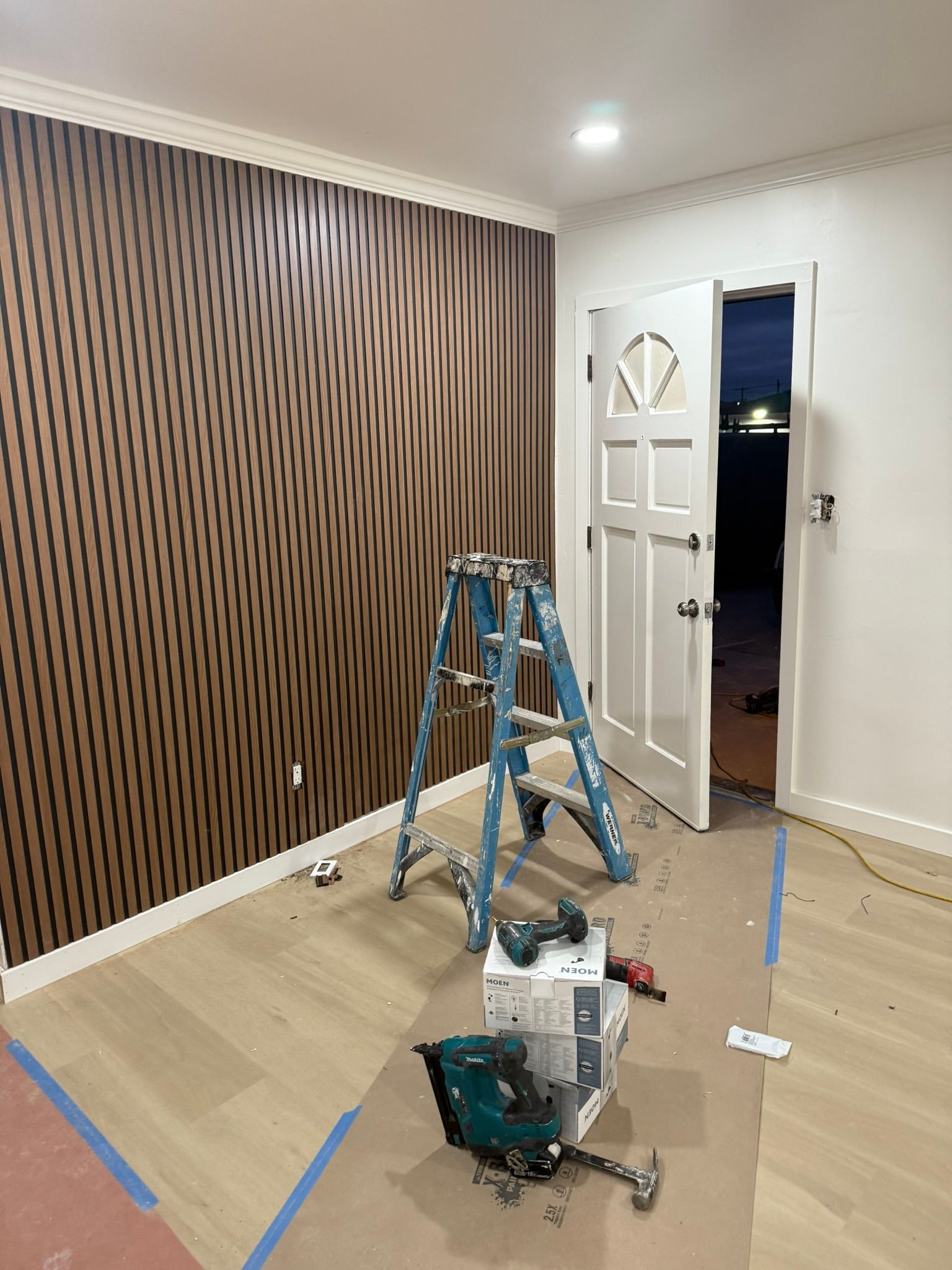 A room under construction with a decorative brown-and-black accent wall, a blue stepladder, tools, and an open doorway.