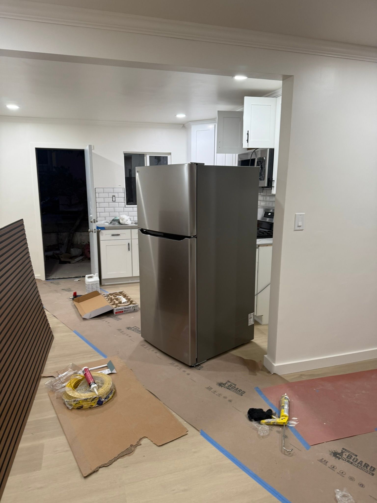 A stainless steel refrigerator sits in the middle of a room under renovation with exposed flooring and construction debris.