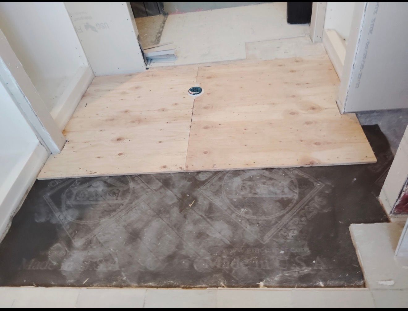Bathroom floor in progress; plywood laid over dark underlayment, drain visible.