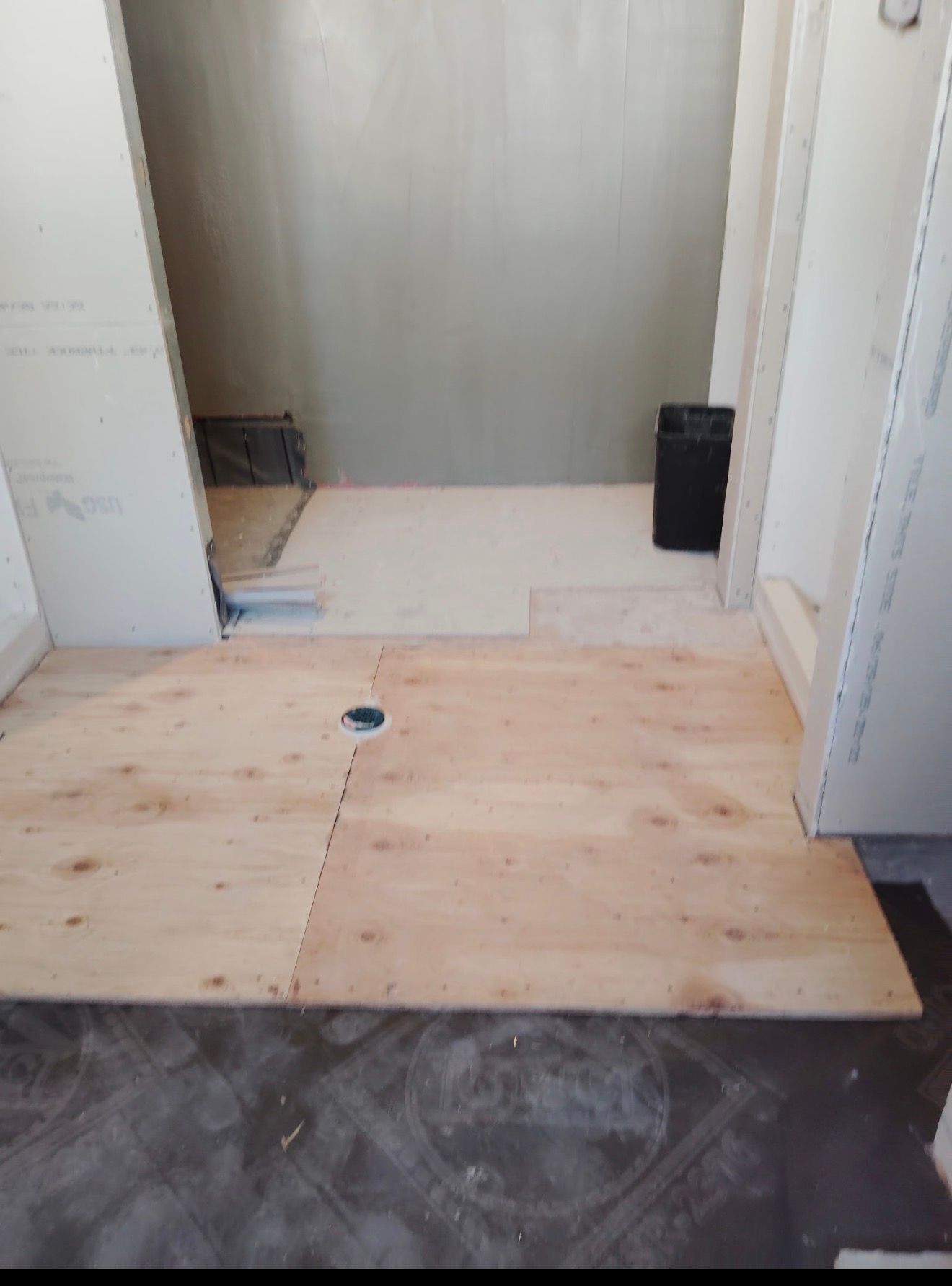 Bathroom under construction, plywood flooring laid, plumbing drain visible. Walls are unfinished, with a small shower area.