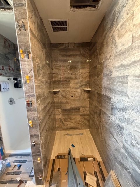 Shower stall with gray tile walls and floor. Two built-in shelves. Floor is partially tiled.