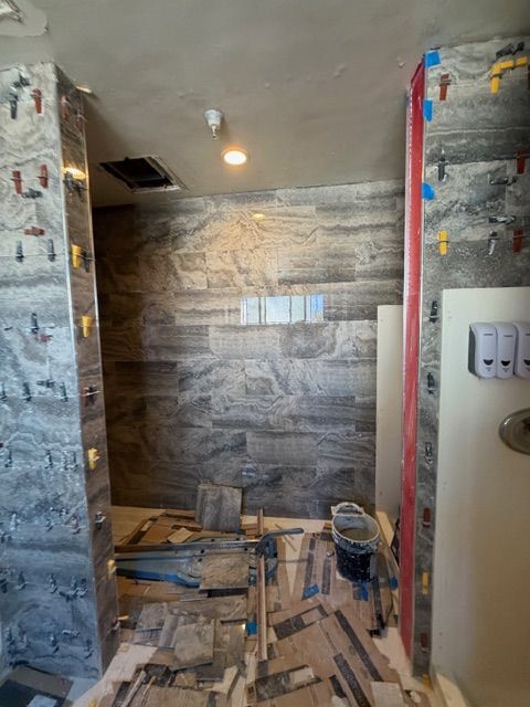 Bathroom under construction, tiling in progress. Gray and brown stone-look tile on walls, floor partially tiled.