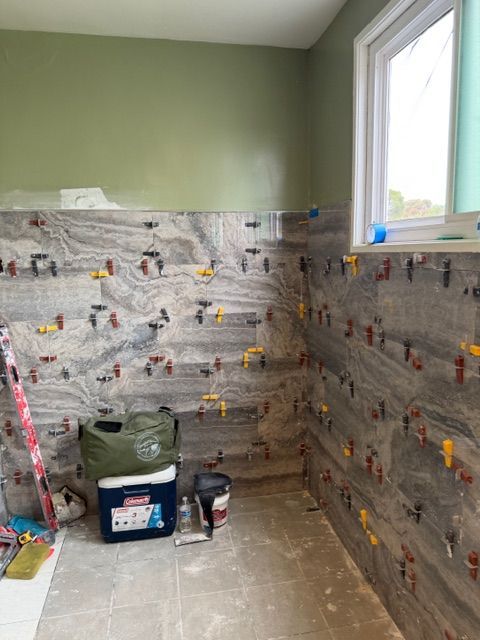 Bathroom with gray and white tiled walls, in process of being installed. Tools and supplies on floor. Window on the right.