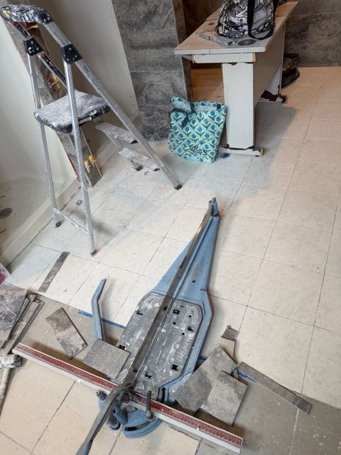 Tile cutter and ladder on a tiled floor during construction; a teal bag and desk are in the background.
