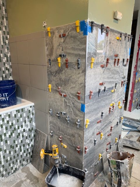 Bathroom wall with large gray tiles being installed, using leveling clips. A drill and bucket are visible.