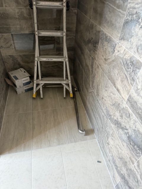 Shower under construction, with a ladder and drain. Grey tiled walls and floor.