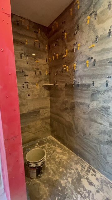 A newly tiled shower with grey stone tiles, leveling clips, and a built-in shelf; bucket of mortar on floor.