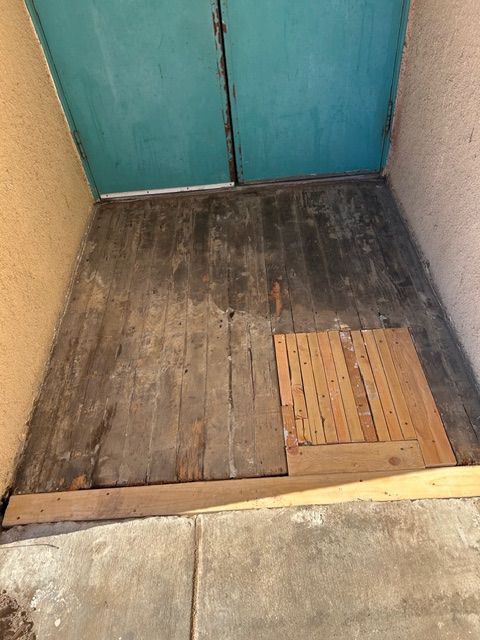 Damaged wooden floor in a doorway. A patch of new wood sits on the floor. Teal doors in the background.