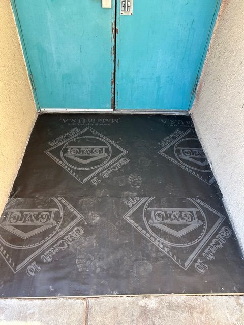 Black mat with gray logo in front of teal double doors. Entrance to a building.