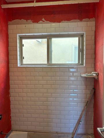 A bathroom wall under construction, partially covered in white subway tiles with red waterproof sealant above and sides.