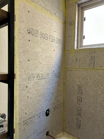 Cement backer board panels installed on shower walls around a small window and plumbing pipes in a residential bathroom.