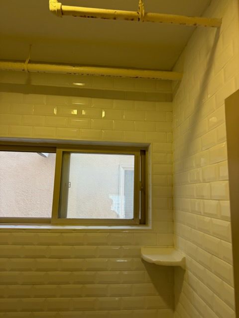 A corner of a tiled bathroom with a window, a corner shelf, and visible yellow pipes mounted on the ceiling.